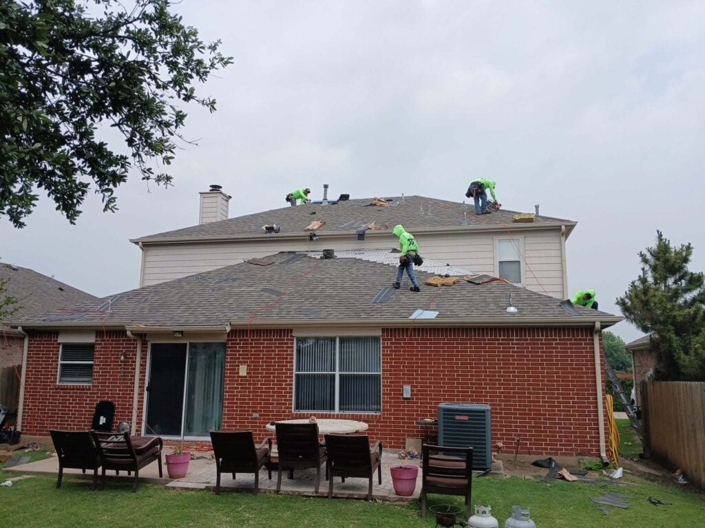 Several roofers in bright green shirts are actively working on the asphalt shingle roof of a two-story brick residential house, with some old shingles removed and new underlayment visible. The backyard patio with furniture and a grassy area is in the foreground.