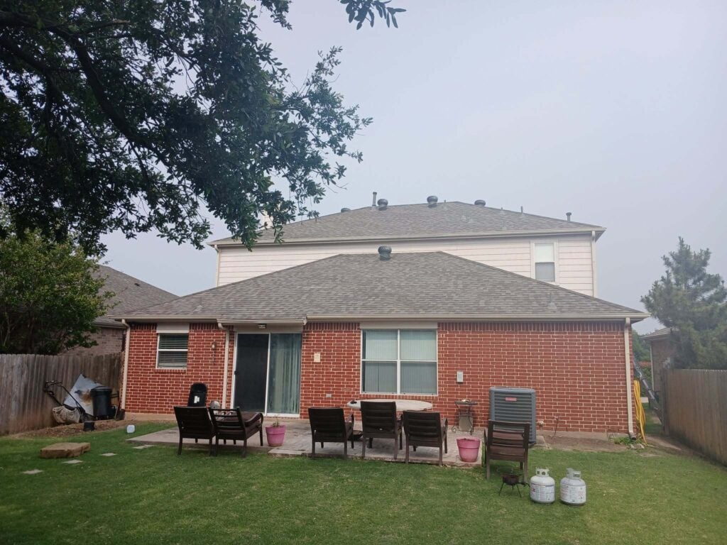 This image shows the back of a two-story residential house with a gray asphalt shingle roof, a brick first story, and light-colored siding on the second story. A patio with outdoor furniture and a grassy backyard are visible in the foreground, with a large tree partially obscuring the upper left.