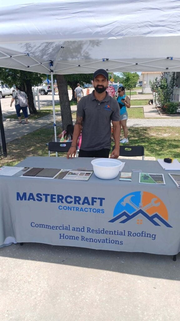 A man wearing a Cedar Crest Roofing polo shirt stands behind a branded booth table displaying roofing material samples and company information at an outdoor event.