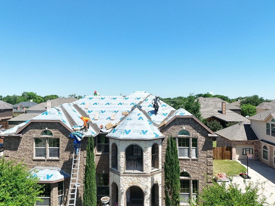An aerial view of a large brick house with a complex roof structure, where several roofers are actively working on installing blue synthetic underlayment. One roofer is visible on a ladder, with a clear blue sky overhead.