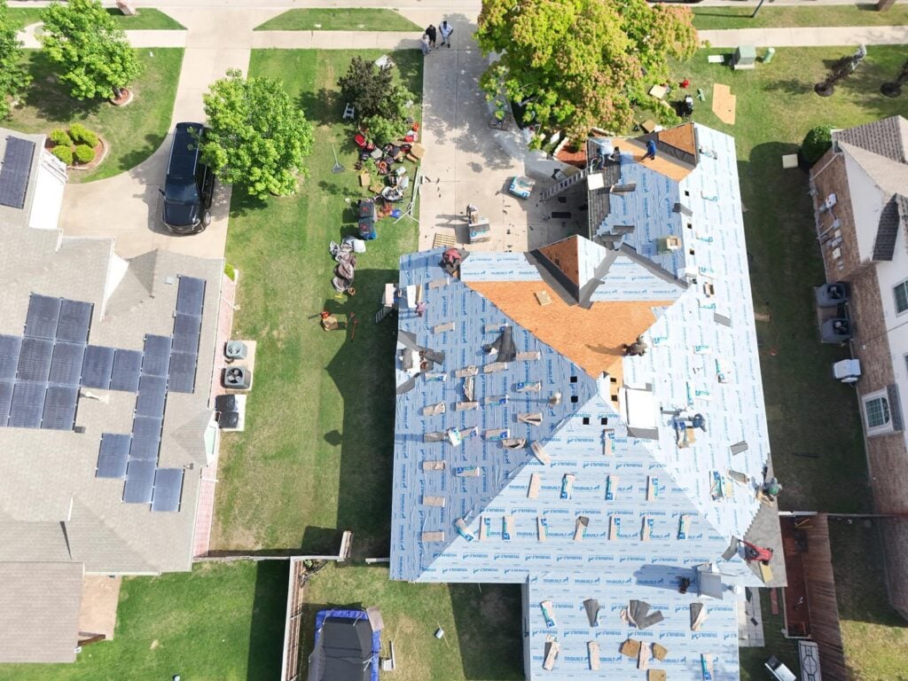 An aerial view of a residential roof undergoing replacement, with blue underlayment installed and bundles of asphalt shingles being laid. Several crew members are visible working on different sections of the roof.