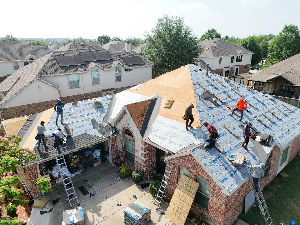 An aerial view of a residential house undergoing a full roof replacement, with multiple crew members actively working on installing underlayment and new asphalt shingles. The roof is partially stripped to the plywood deck, and ladders and material bundles are visible around the property.