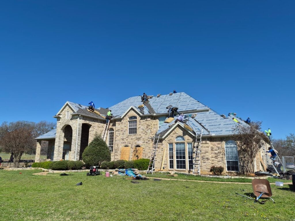 A large brick house with a complex roofline is shown with multiple roofers actively working, installing underlayment and new shingles. Ladders are positioned around the house, and roofing materials are visible on the ground.