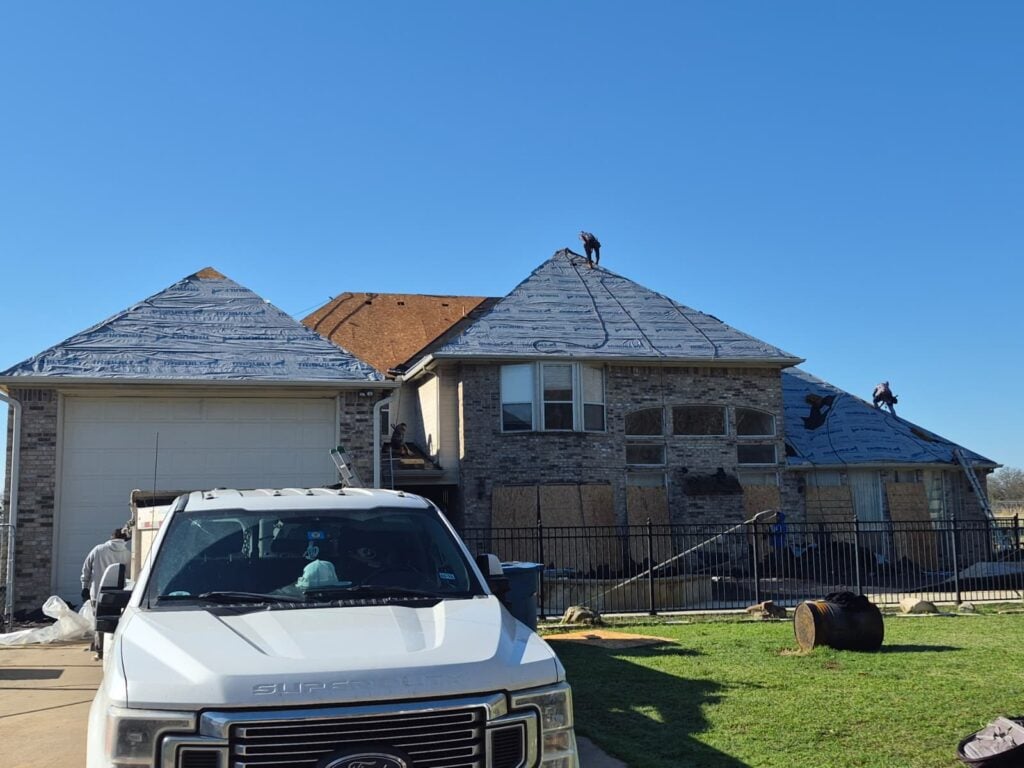 This image shows a large residential house undergoing roof replacement, with blue synthetic underlayment installed on most sections and workers actively on the roof. A white Ford Super Duty truck is parked in the driveway, partially obscuring the garage.