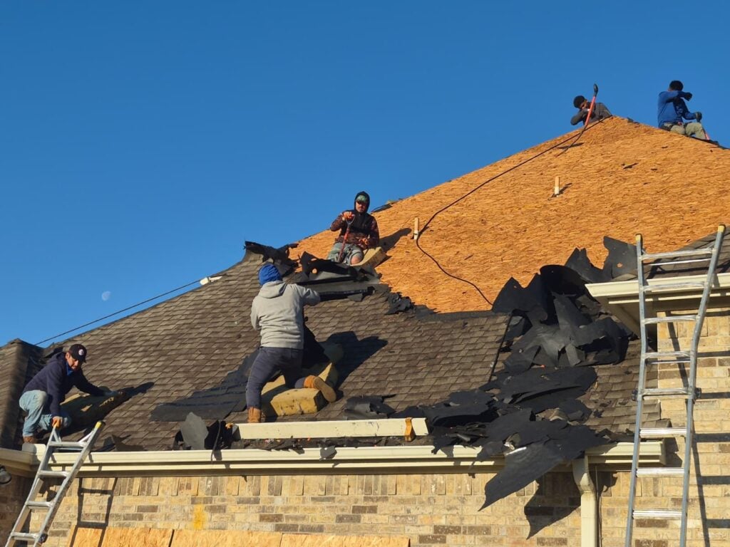Several roofers are actively removing old asphalt shingles from a residential roof, exposing the plywood decking underneath. A ladder is visible on the right, and the sky is clear blue with a faint moon.