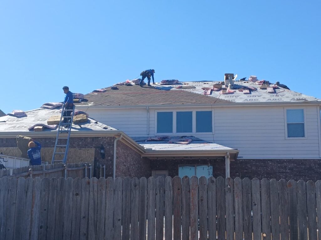 Roofers are actively working on replacing the asphalt shingle roof of a residential house, with bundles of new shingles and underlayment visible on the roof deck.
