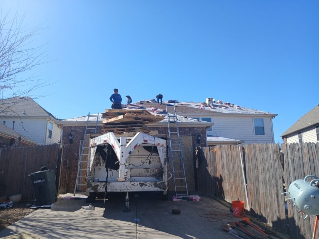Several roofers are actively working on a residential roof, which is partially stripped with new bundles of shingles visible. A large trailer and ladders are positioned in the driveway, with a clear blue sky overhead.