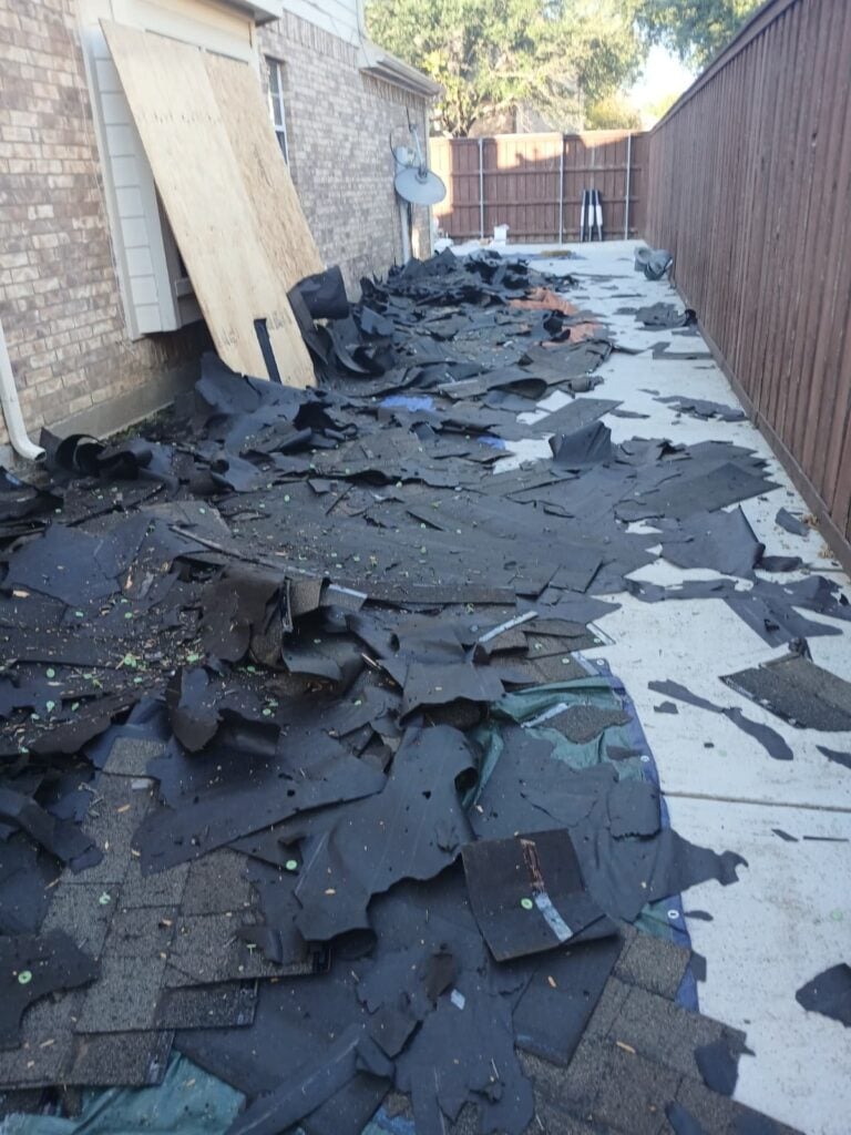 A narrow side yard is completely covered with discarded asphalt shingles and underlayment, indicating a roof tear-off in progress. A large sheet of plywood leans against the brick wall of the house.