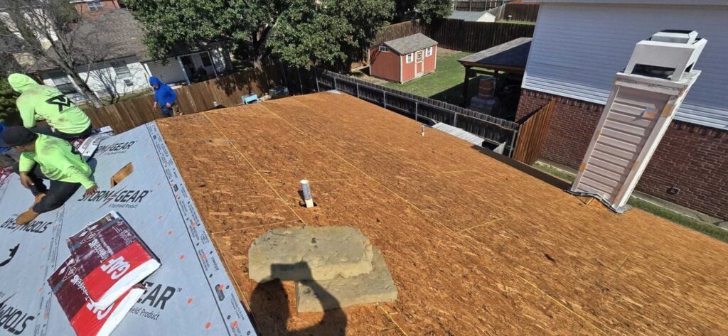 Roofers are installing underlayment on a residential roof, with two crew members visible in the foreground and one further back. The image shows the exposed roof deck, a chimney, and a vent pipe, with a backyard and shed in the background.