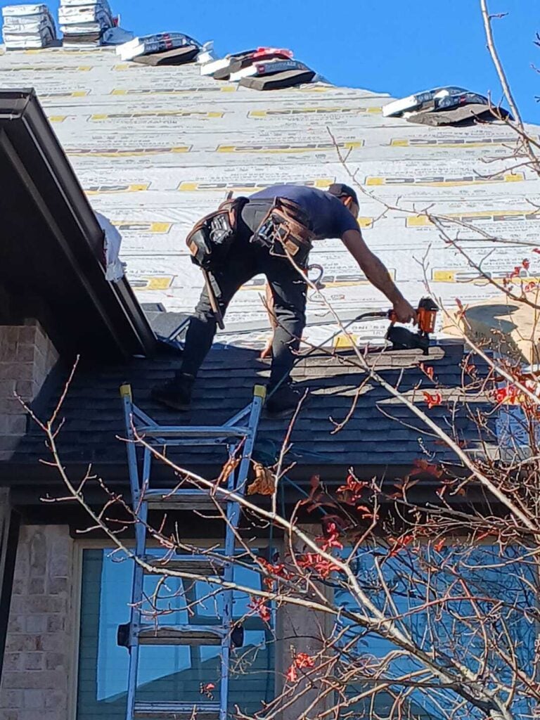 A roofer is actively installing asphalt shingles on a residential roof, with underlayment visible on the upper sections. Bundles of new shingles are stacked on the roof, and a ladder is positioned against the lower roofline.