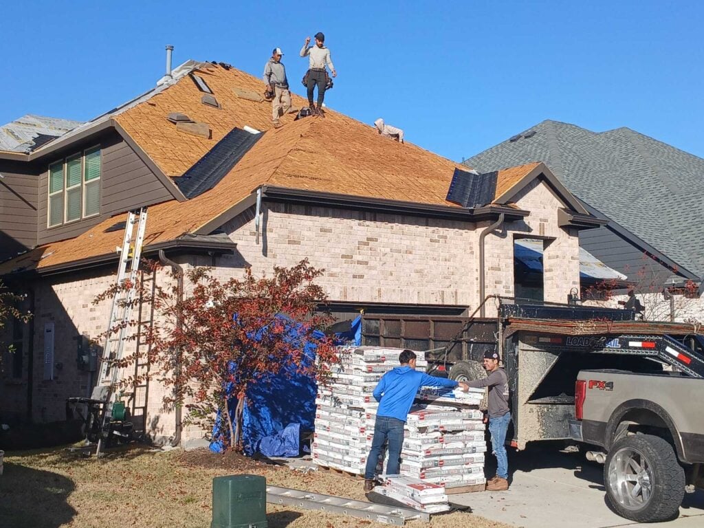 Roofing crew actively working on a residential roof, with some workers on the roof and others unloading pallets of asphalt shingles from a truck and trailer on the ground.