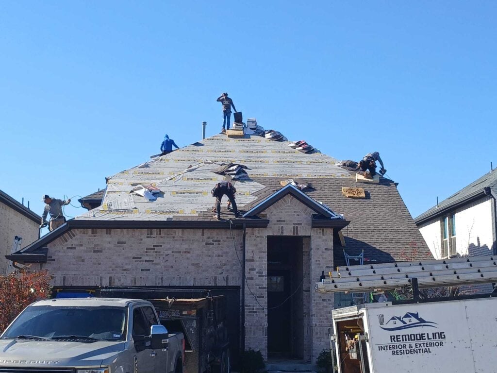 Several roofers are actively working on a residential roof, which is partially stripped and has new underlayment installed. Bundles of new asphalt shingles are visible on the roof, and a work truck is parked in the driveway.