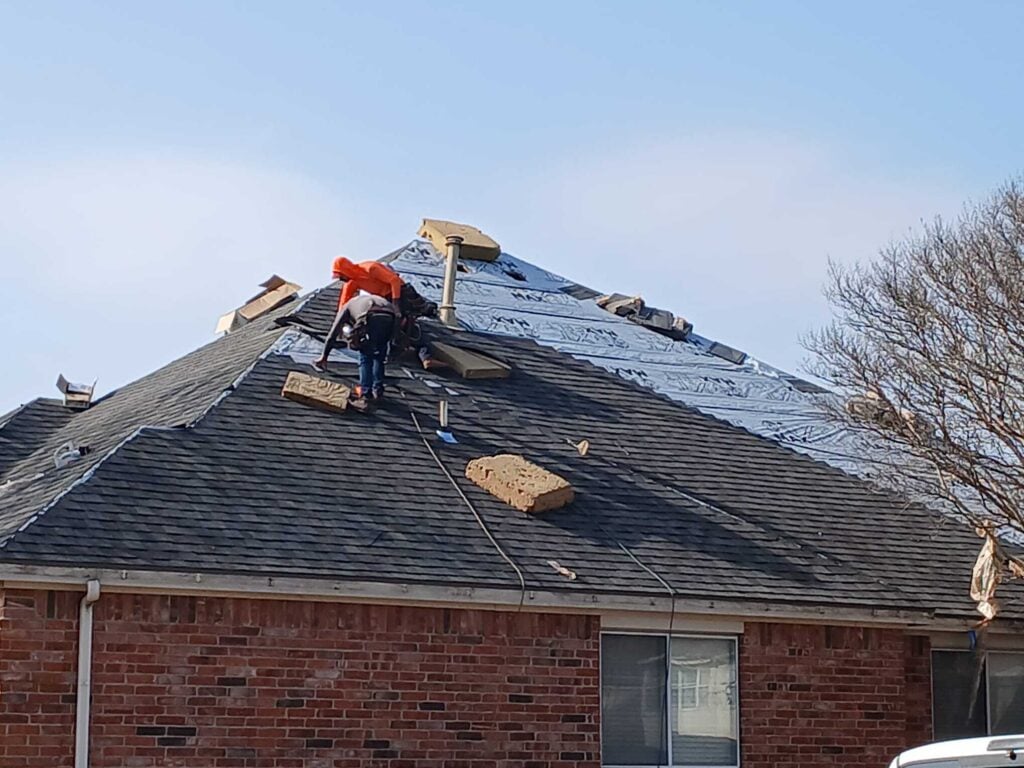 Two roofers are actively working on a residential roof, with one section covered in blue underlayment and another section already shingled with dark asphalt shingles.
